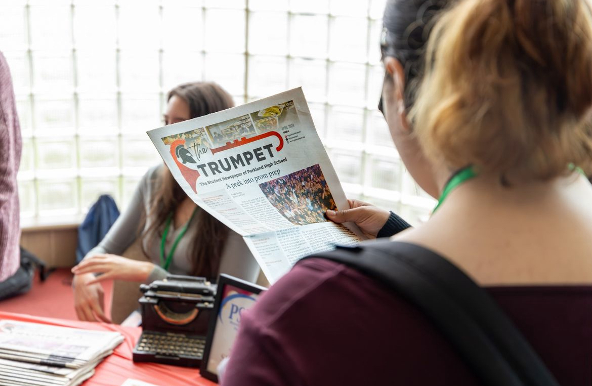 A student admiring The Trumpet newspaper at its table while Editor-in-Chief Sarah George networks in the background. 