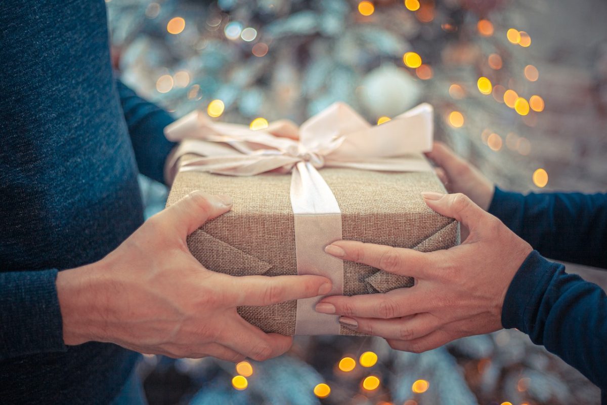 Two sets of hands exchanging a present wrapped in tan paper infront of a festively decorated tree.